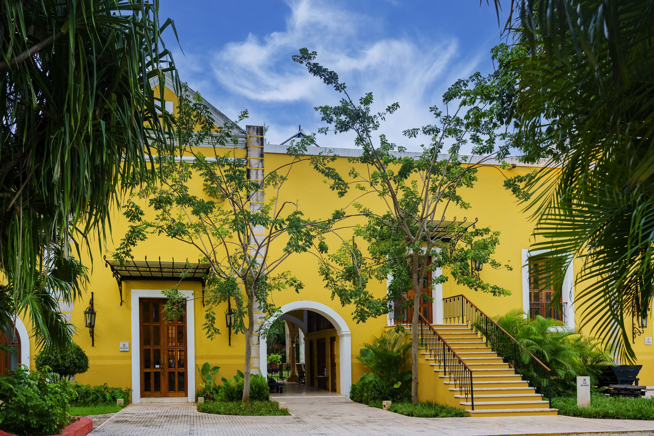 Bright yellow building with stairs surrounded by lush greenery at Hacienda Xcanatun, Angsana Heritage Collection.